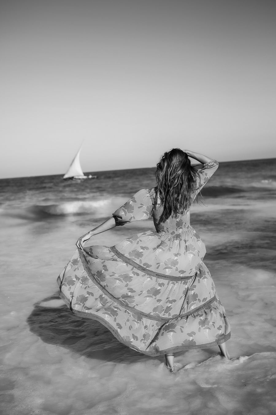 This is a black and white photo of a women wearing a long flowy dress at the beach.