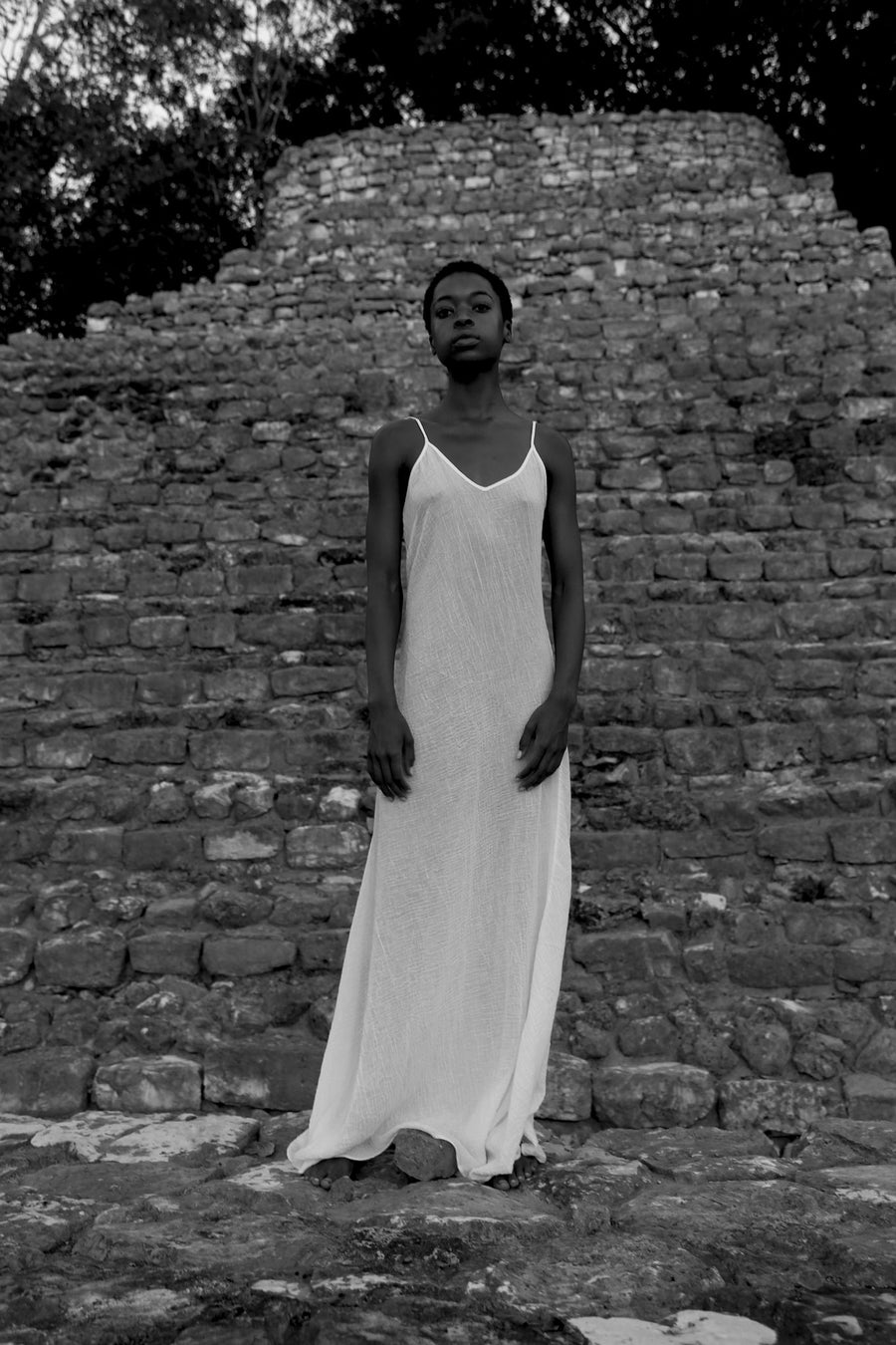 This is a black and white photo of a womand in a white slip dress syanding in front of a monument made of stones.