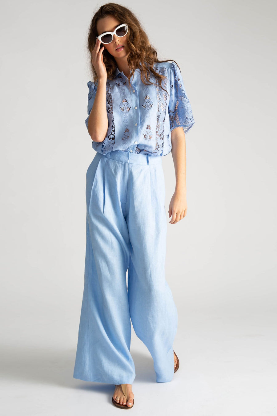 This is a photo of a woman wearing an all blue outfit. Her top is linen with lace embroidery with a button down front, linen collar, and short sleeves. The shirt is tucked into a pair of loose-fit solid blue linen pants with belt loops and side seam pockets. She styles the outfit with white sunglasses and nude sandals.