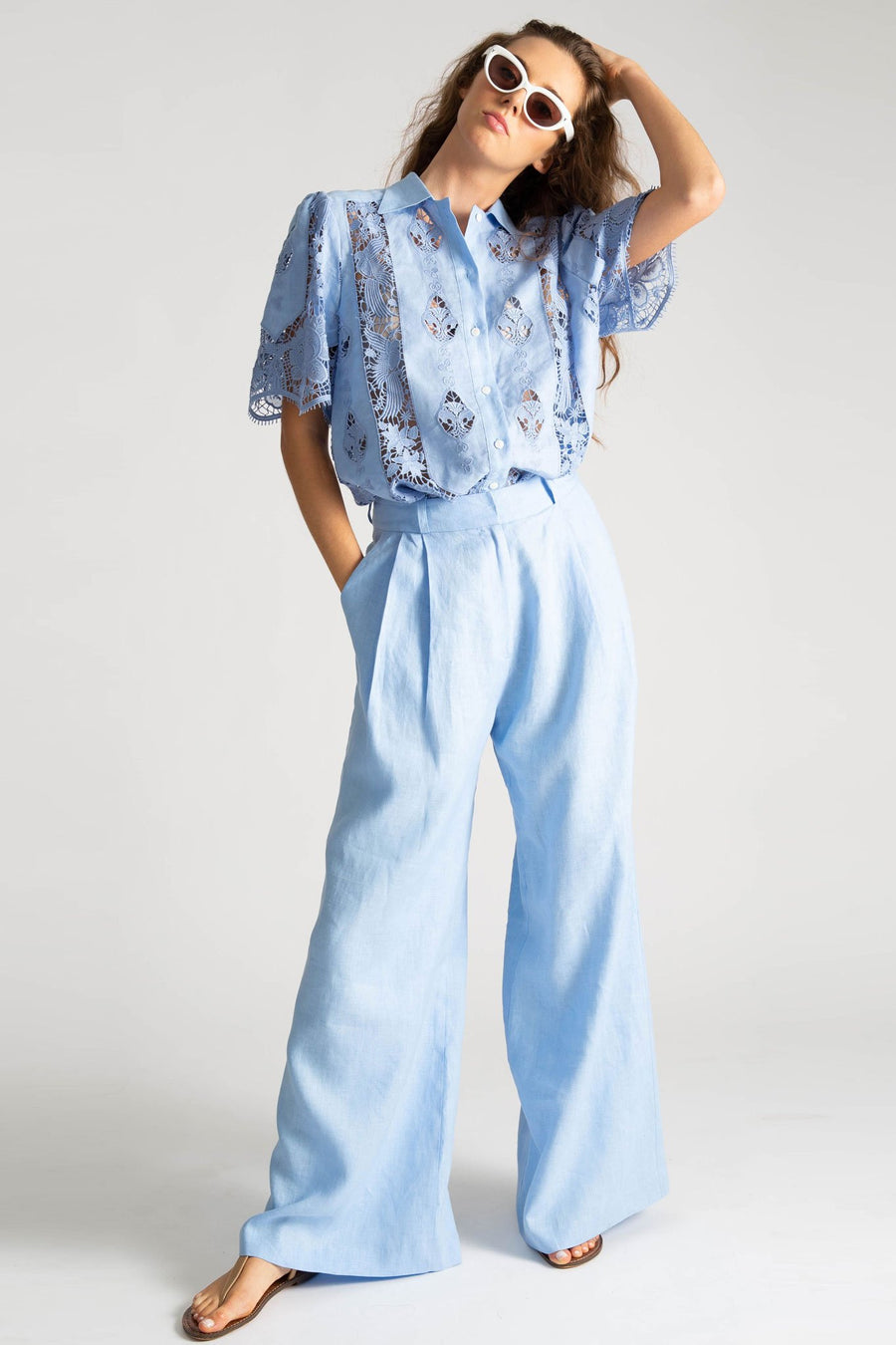 This is a photo of a woman wearing an all blue outfit. Her top is linen with lace embroidery with a button down front, linen collar, and short sleeves. The shirt is tucked into a pair of loose-fit solid blue linen pants with belt loops and side seam pockets. She styles the outfit with white sunglasses and nude sandals.