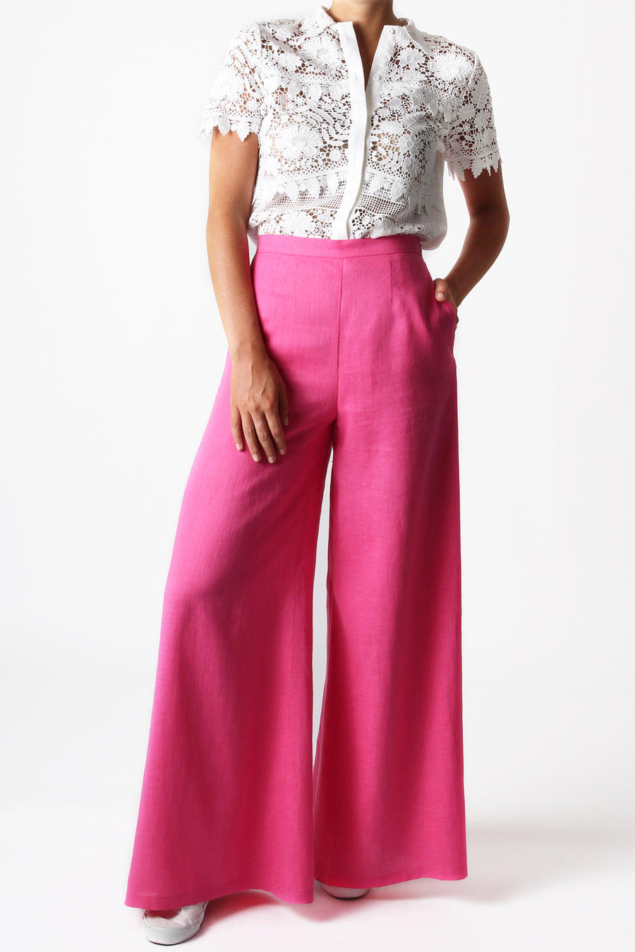 This is a photo of a woman from the neck down, she is wearing a button front, lace tired blouse tucked into a pair of bright pink, flare leg pants. She has her hand in the left side pocket of the pants.
