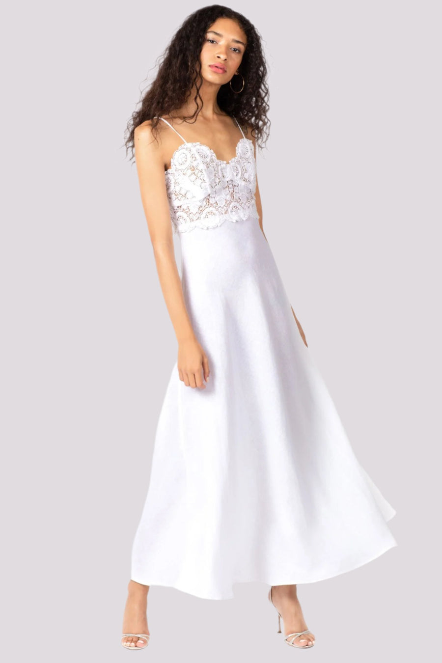 This is a photo of a woman wearing a floor length, pure white, linen and lace dress. The dress has spaghetti straps and a fully lace bodice, with a flowing linen skirt.