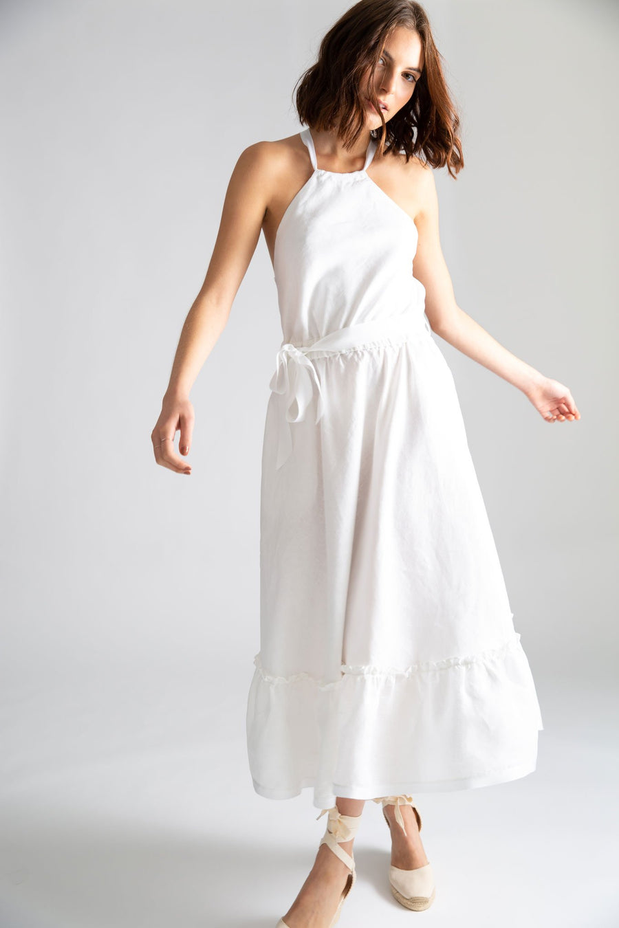 This is a photo of a woman wearing a white linen dress with halter neckline, waistline elastic and removable gross grain ribbon. It has a bottom ruffle detail that falls just above the ankle and is finished with a gross grain plain white ribbon.