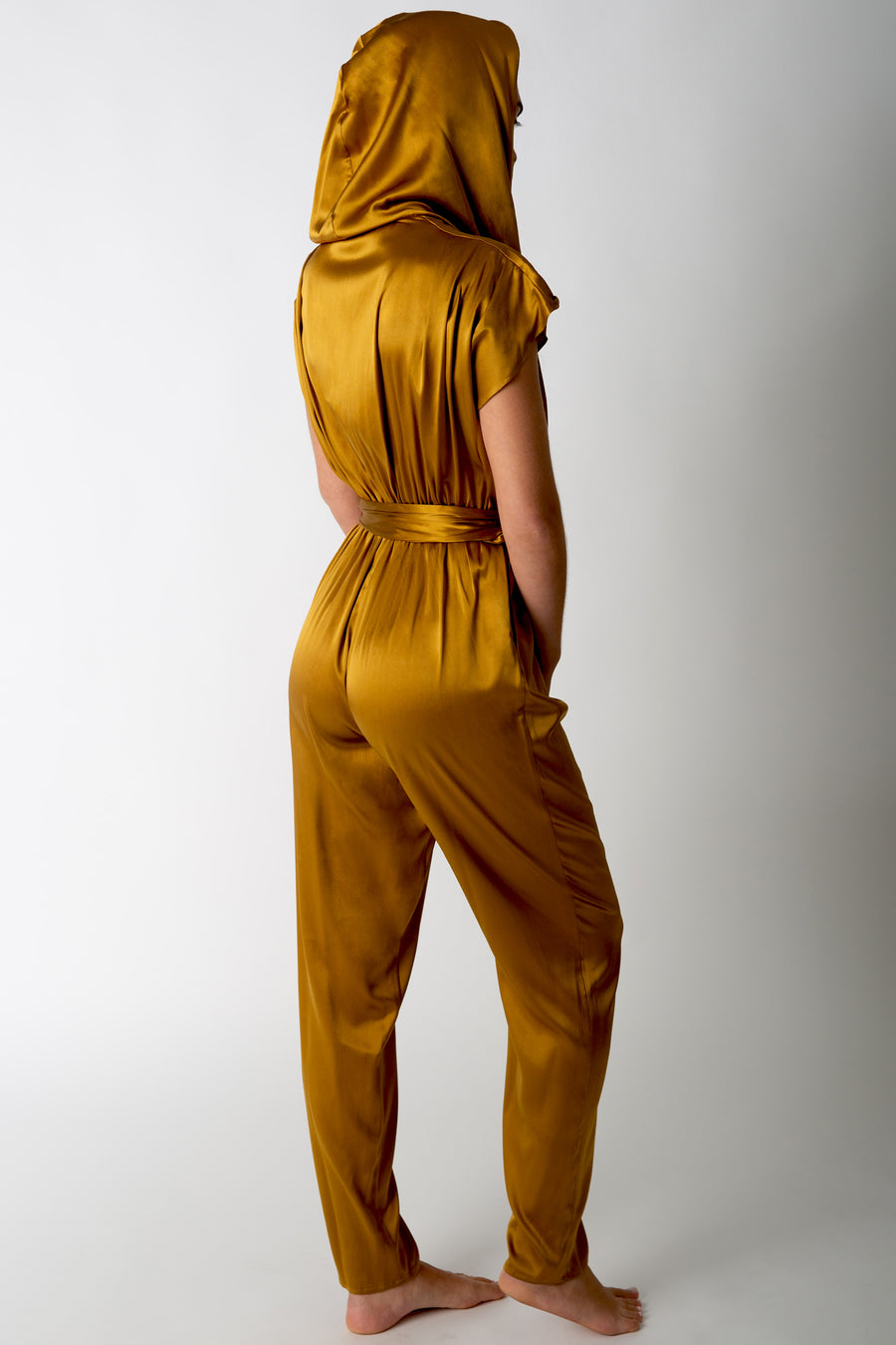 This is a back view of a woman wearing a silk gold jumpsuit with hood up.