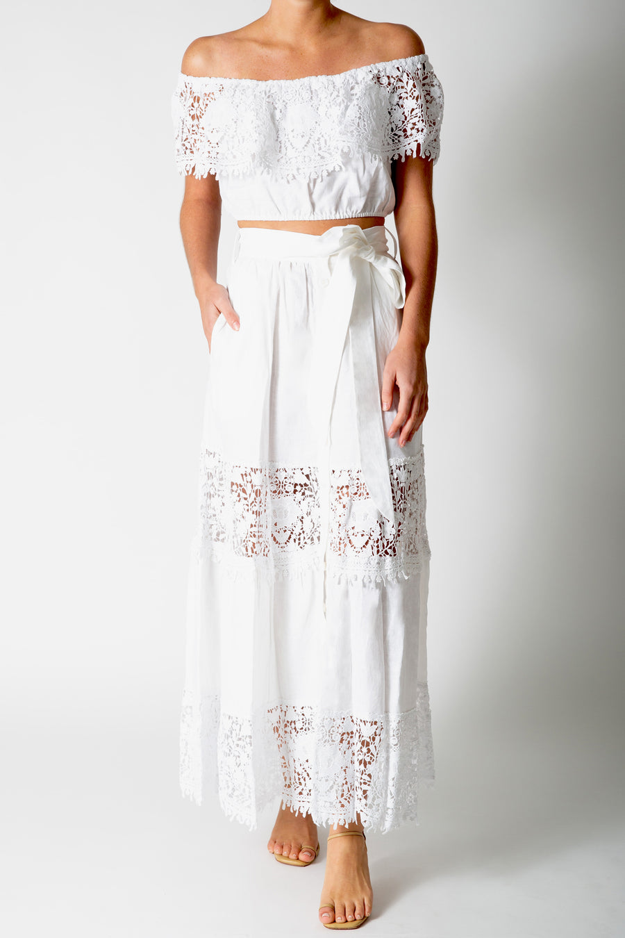 This is a photo of a woman wearing a white lace crop top with a matching white maxi skirt. She stands with her right hand in the skirt pocket.