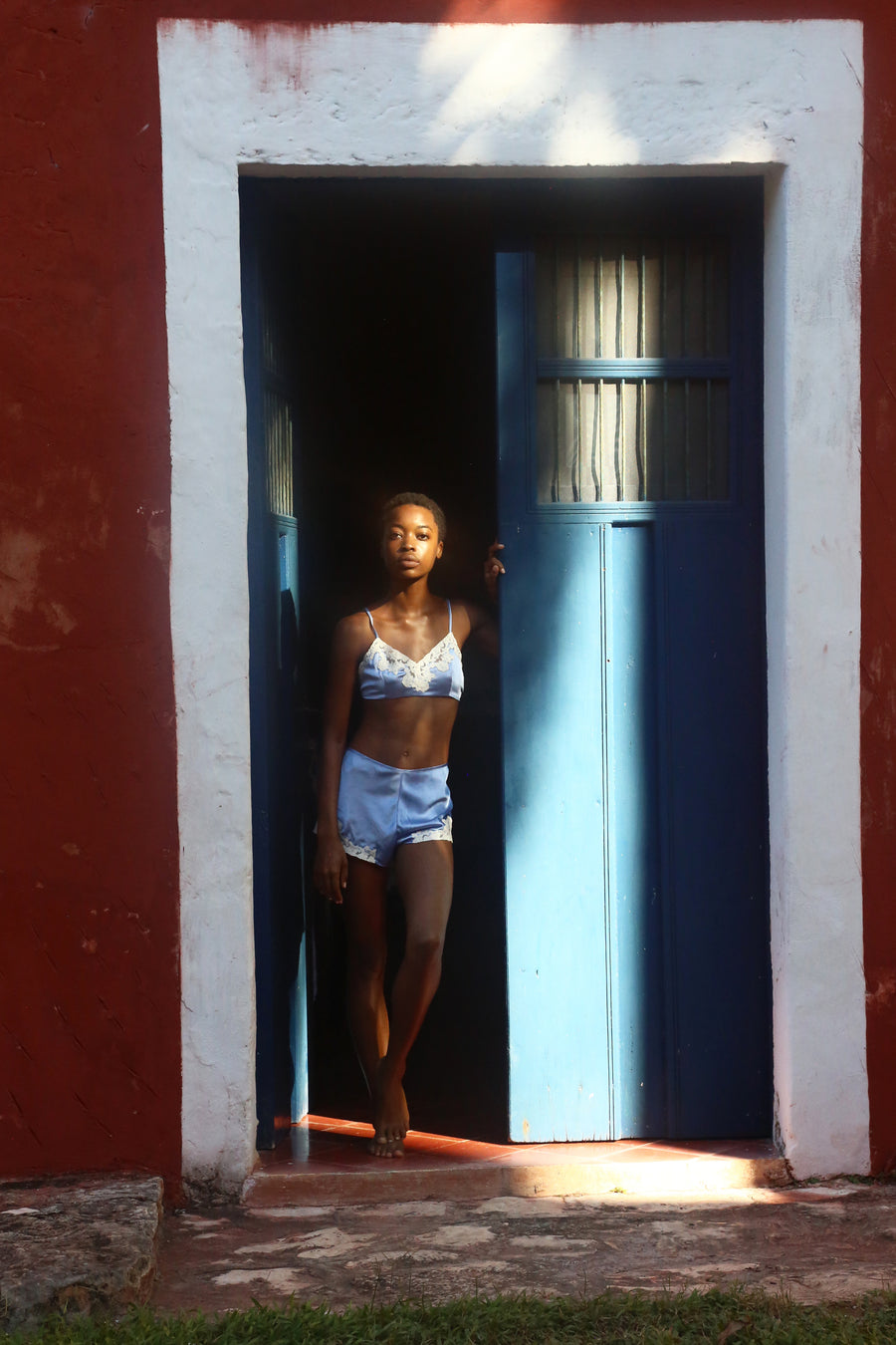This is a photo of a woman wearing a periwinkle colored silk set of mini shorts and bralette with lace trim. She stands in the doorway of a rustic building with red walls and a blue door.