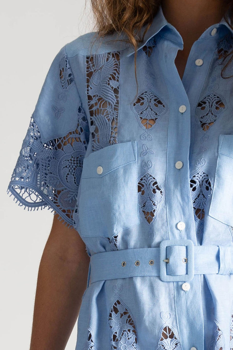 This is a detail photo of a woman wearing a light blue linen and lace dress with linen collar detail and button up front center for closure. The dress is shown with a matching linen belt that can be removed. The square pockets shown on the chest have option for closure with small white buttons.