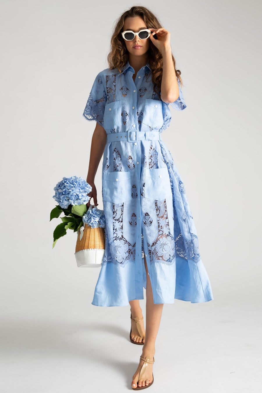 This is a photo of a woman wearing a light blue mid length dress with a button down front, 2 front square pockets on chest, and 2 front square pockets below waistline. The dress is styled with a removable blue linen belt, white sunglasses, and a white and natural straw bag filled with blue hibiscus flowers.