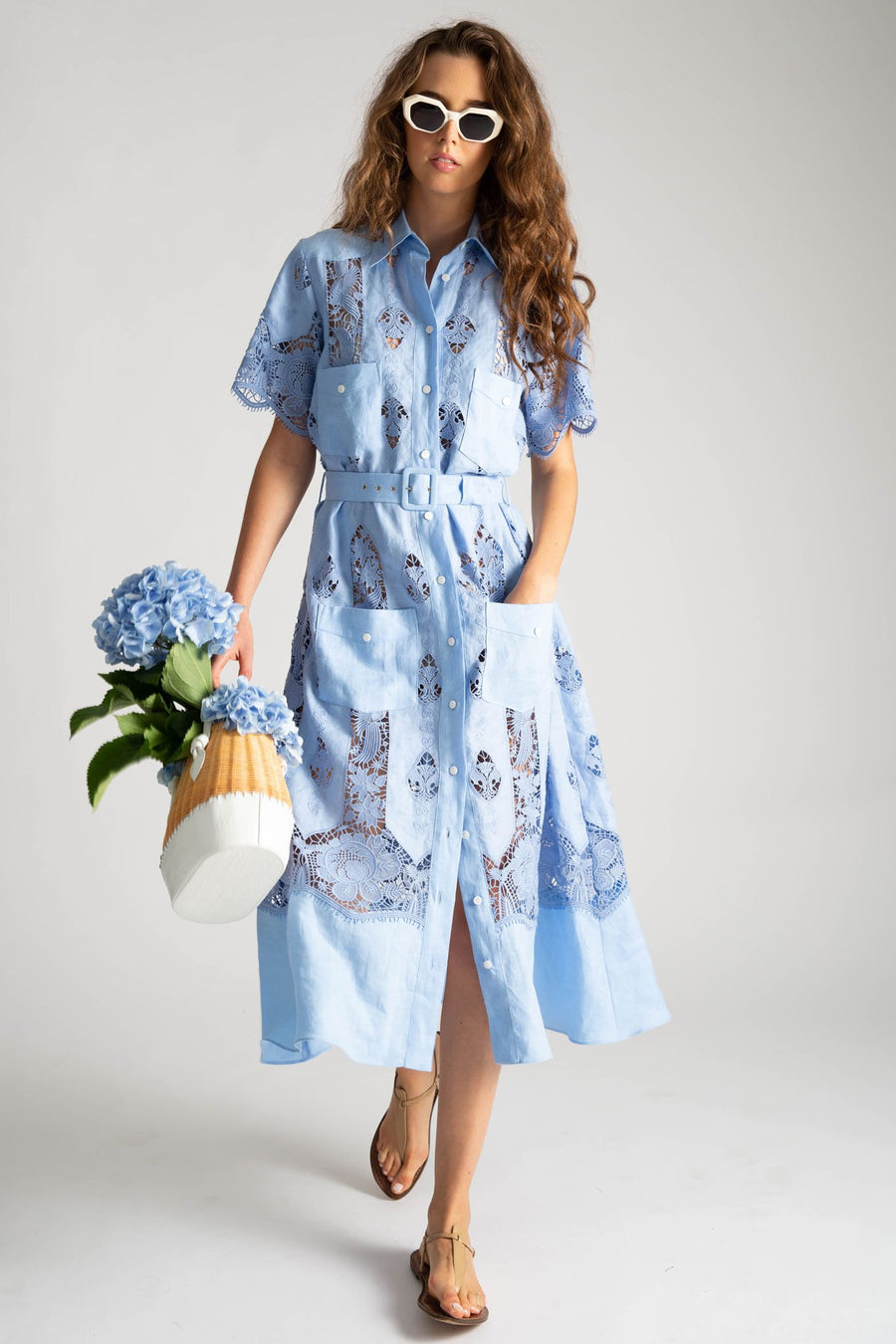 This is a photo of a woman wearing a light blue mid length dress with a button down front, 2 front square pockets on chest, and 2 front square pockets below waistline. The dress is styled with a removable blue linen belt, white sunglasses, and a white and natural straw bag filled with blue hibiscus flowers.
