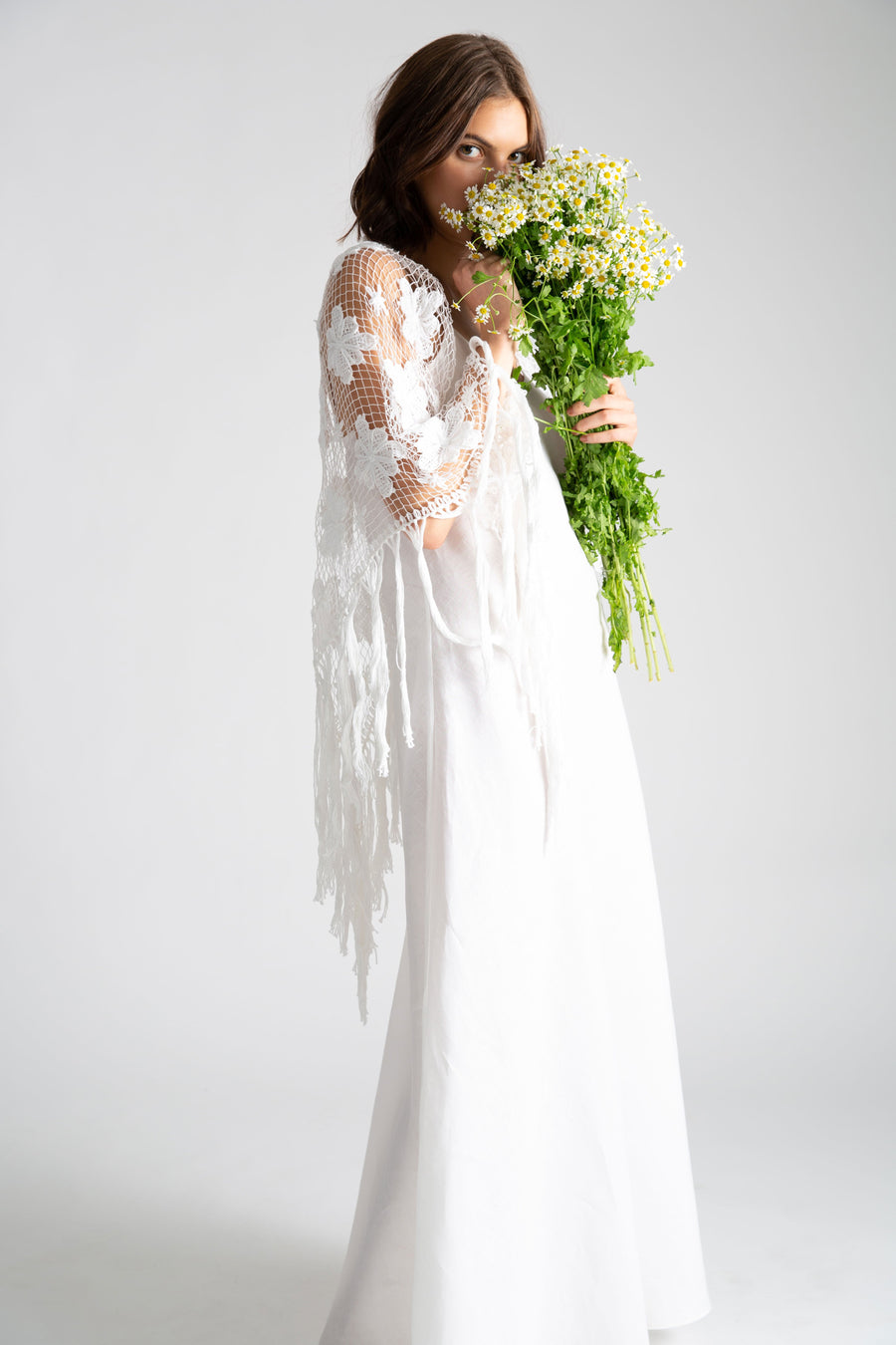 This is a photo of a woman wearing a white linen slip dress with v neck front and side leg slit. Over the slip dress, she wears a white hand knit shawl and holds a bouquet of daisy flowers.