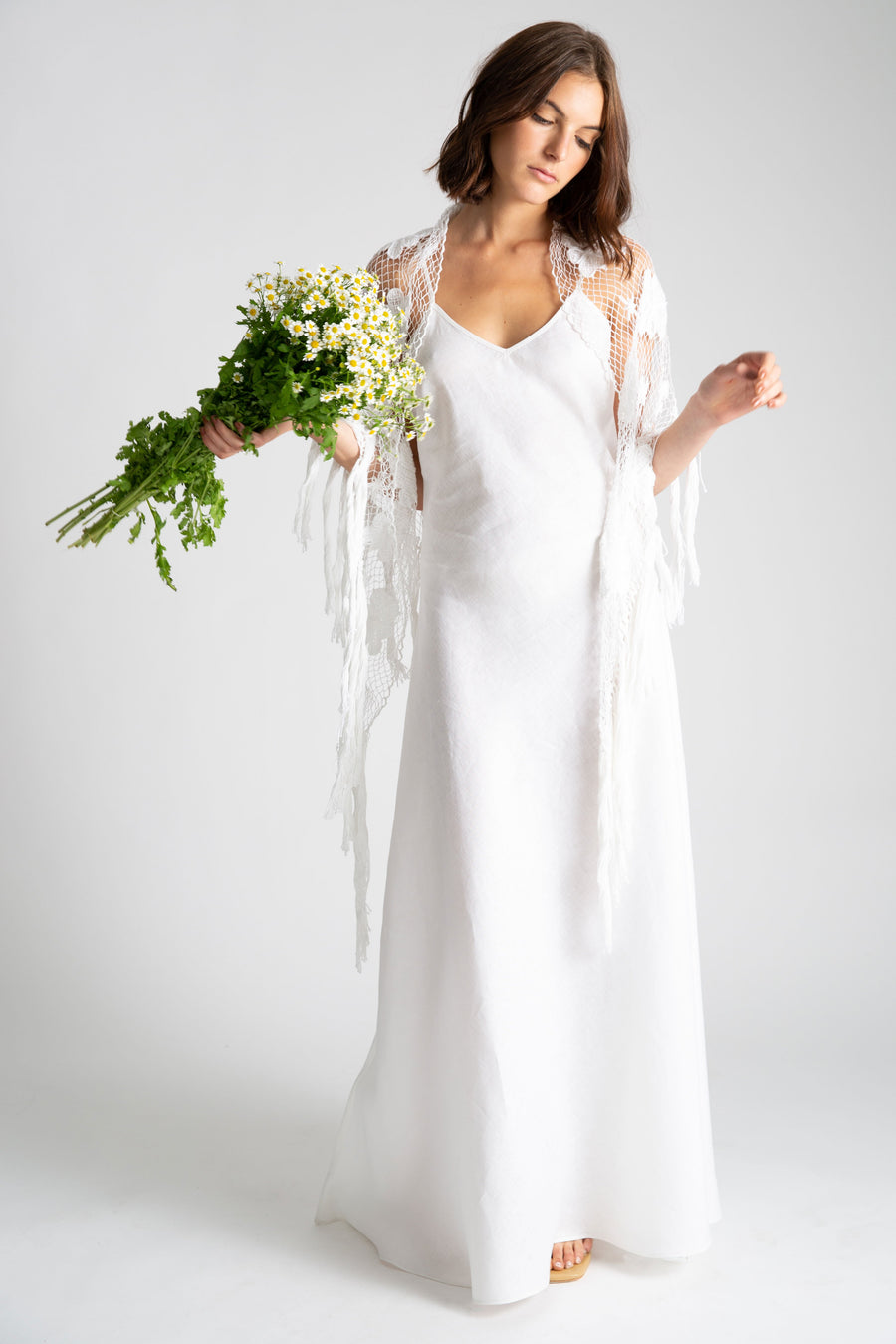 This is a photo of a woman wearing a white linen slip dress with v neck front and side leg slit. Over the slip dress, she wears a white hand knit shawl and holds a bouquet of daisy flowers.