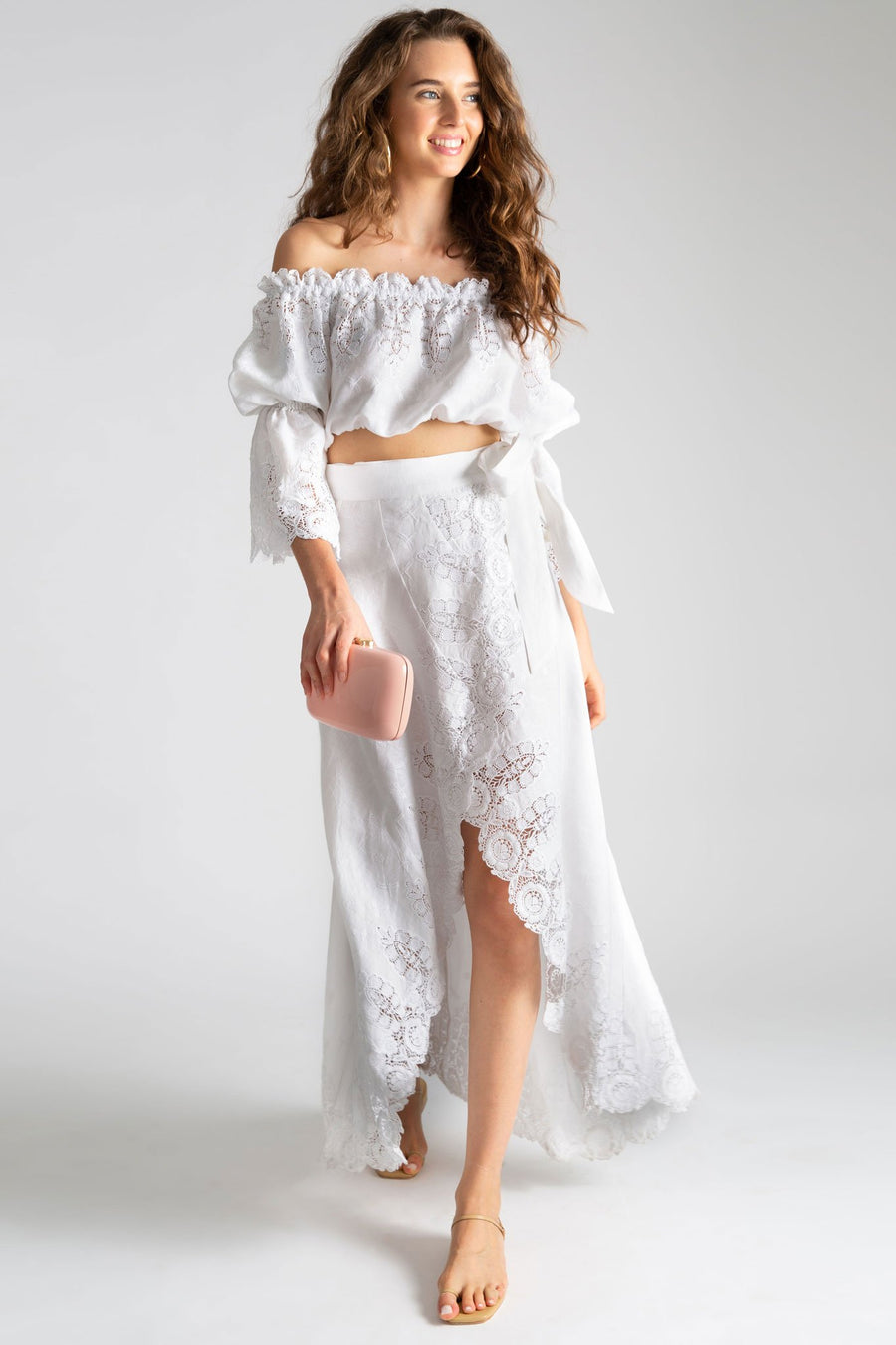 This is a photo of a woman wearing a white linen and lace outfit with maxi wrap skirt open in the front and off the shoulder top with peasant sleeves. She holds a light pink clutch and styles the look with nude sandals.