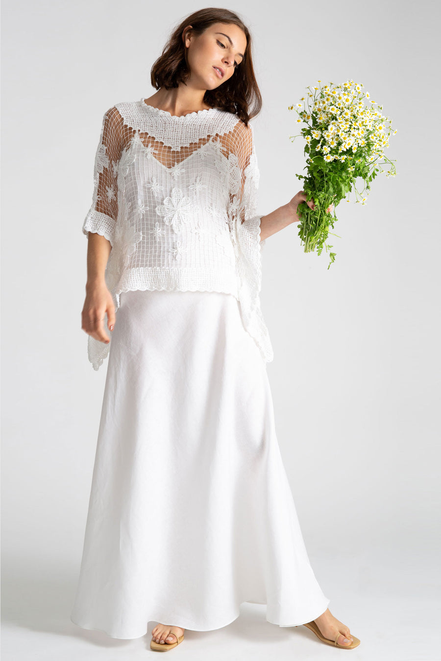 This is a photo of a woman wearing a white linen slip dress with a handmade white crochet knit poncho shawl on top and is holding a bouquet of mini daisies.