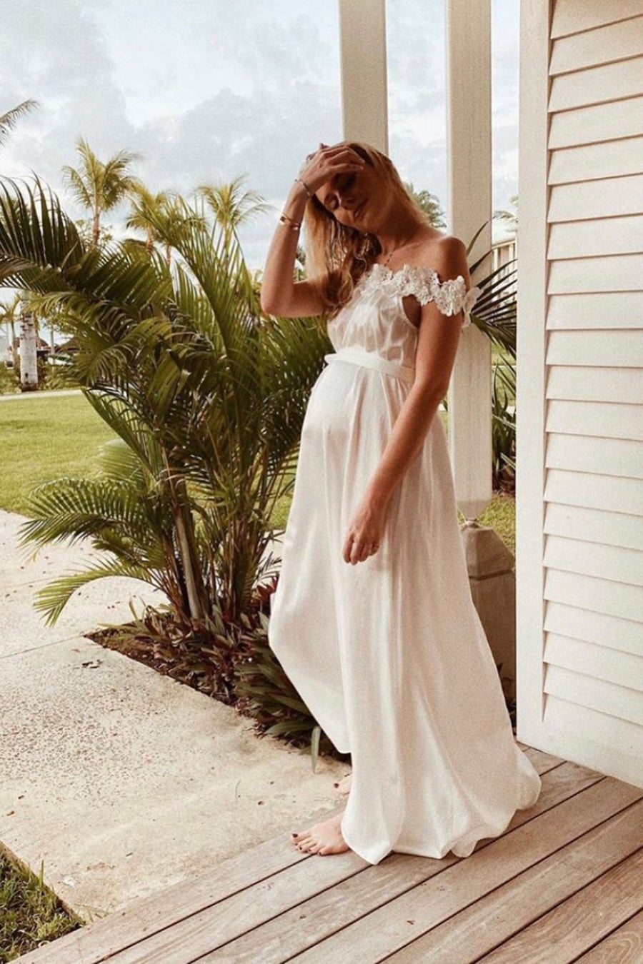 This is a photo of a pregnant woman wearing an off the shoulder, white, floor length dress, that has embroidered flower trim all around the neckline.