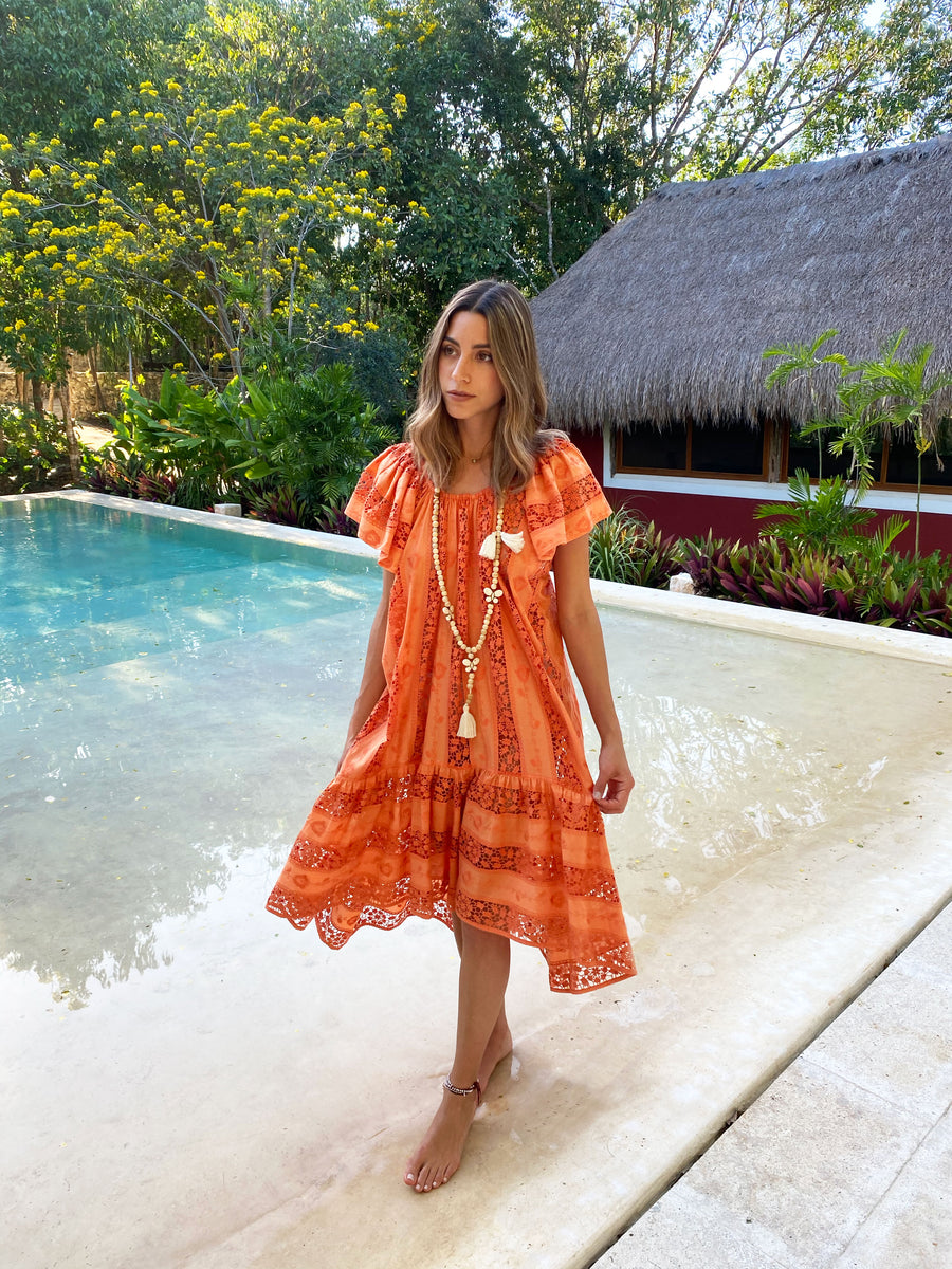 This is a photo of a woman wearing an orange embroidered cover up. She walks alongside of a pool.