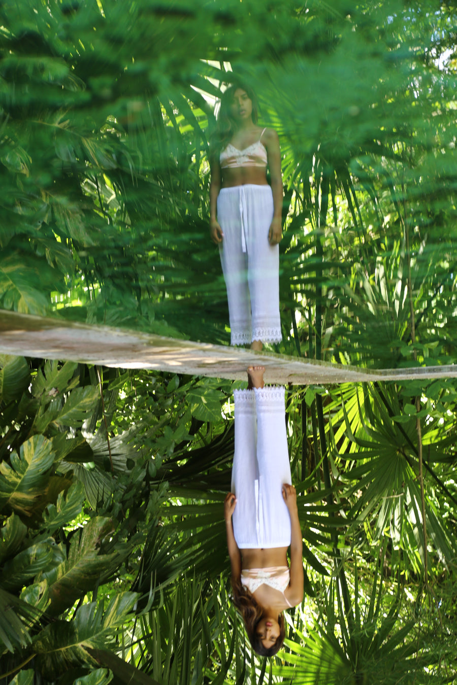 This is an editorial photo of a woman wearing white pants and pink bralette top overlooking her reflection in a pool with green trees behind her.