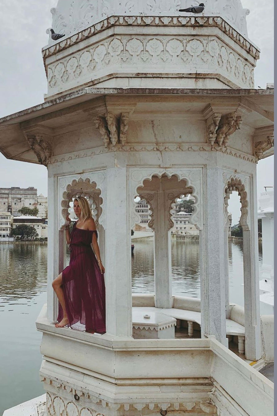 This is a photo of a woman wearing a magenta cotton gauze floor-length maxi coverup that falls off both shoulder and ties around center front chest area and waistline. Photo is taken on a white gazebo type structure overlooking a body of water and dress shows center front leg slit.