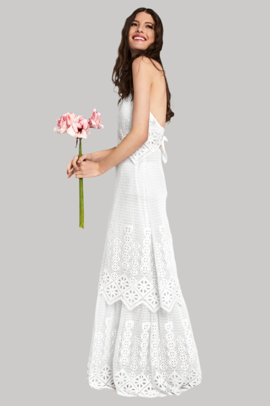 This is a photo of a woman wearing a long white halter dress. She looks over her shoulder and smiles as she holds a bouquet of pink flowers.