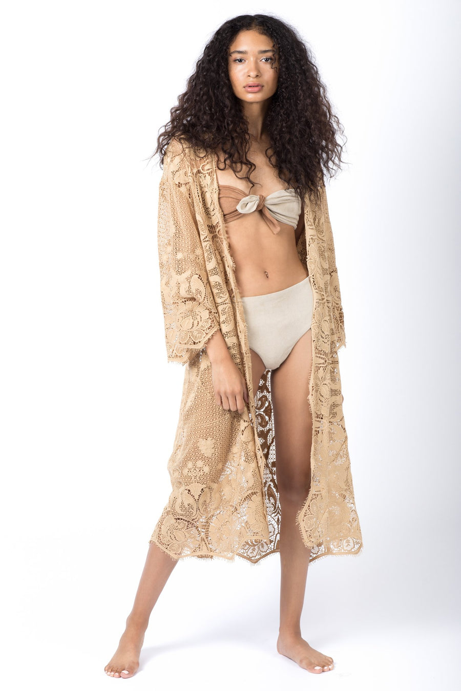 This is a photo of a woman wearing sand-colored high waisted bikini bottoms and a matching bikini top that knots in the middle. Over the bikini, she wears a tan, laced cover up.