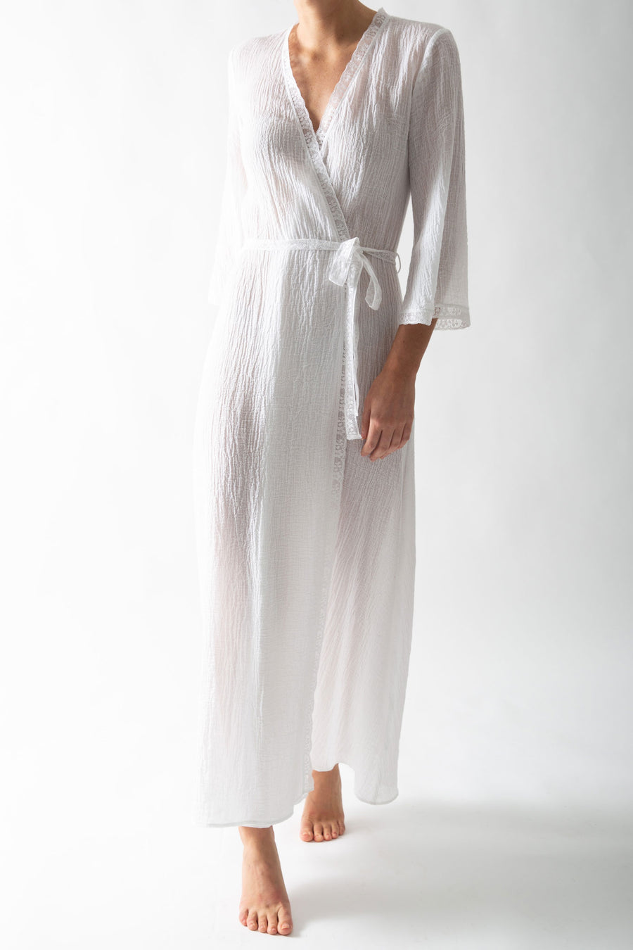 This is a photo of a woman wearing a white cotton gauze rope with wrap closure. There is lace trim along the seams and she wears it belted closed.