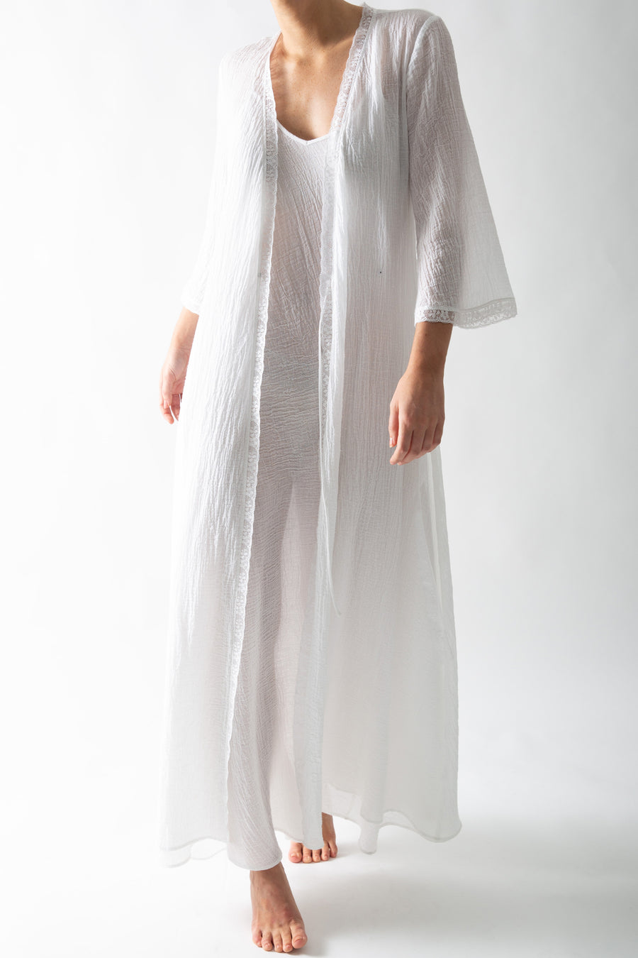 This is a photo of a woman wearing a white cotton gauze rope with wrap closure. There is lace trim along the seams and she wears it open with a white slip dress underneath.