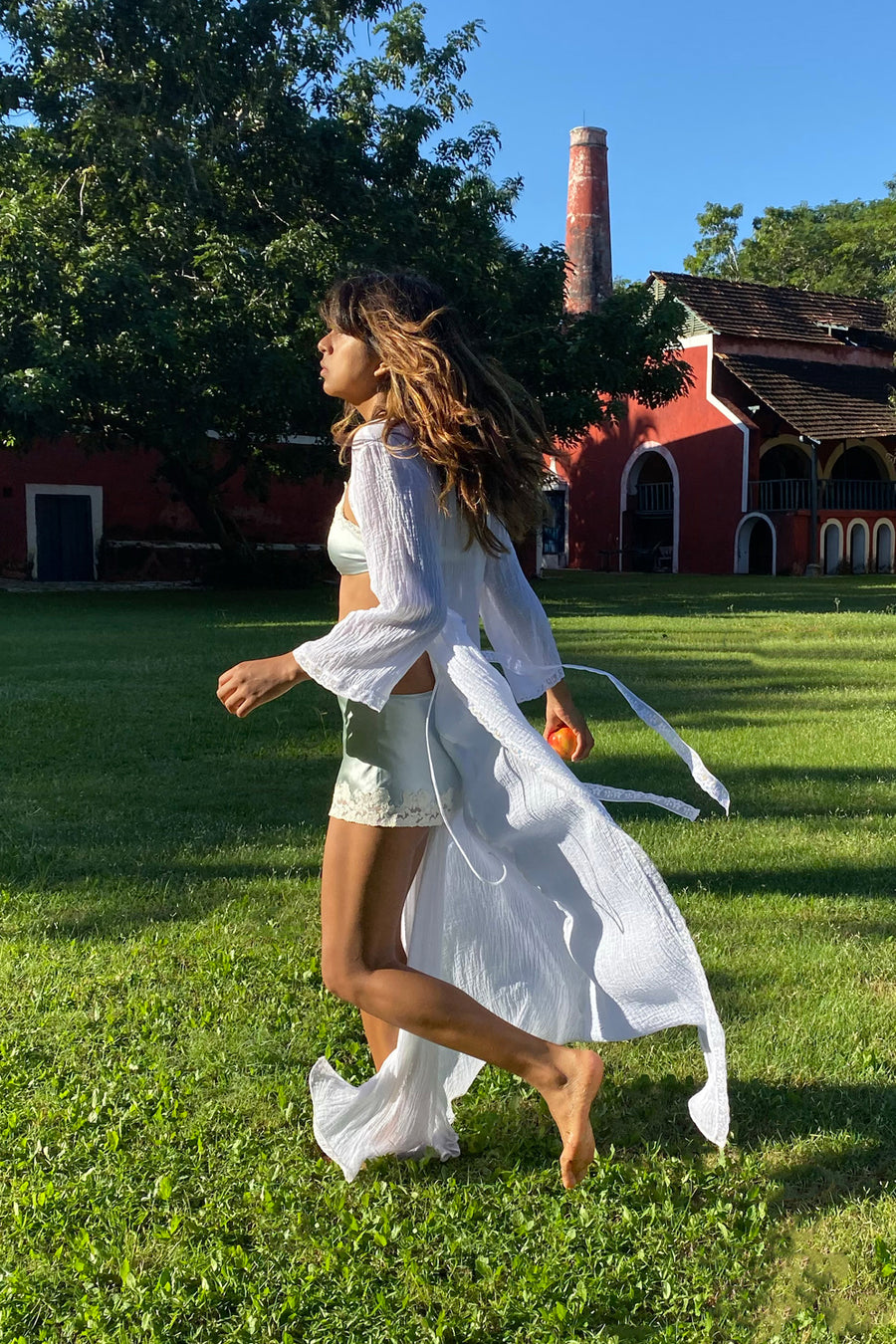 This is a photo of a woman running on grass wearing a seafoam-colored silk set with shorts and bra top. Overtop, she has a white cotton gauze robe.