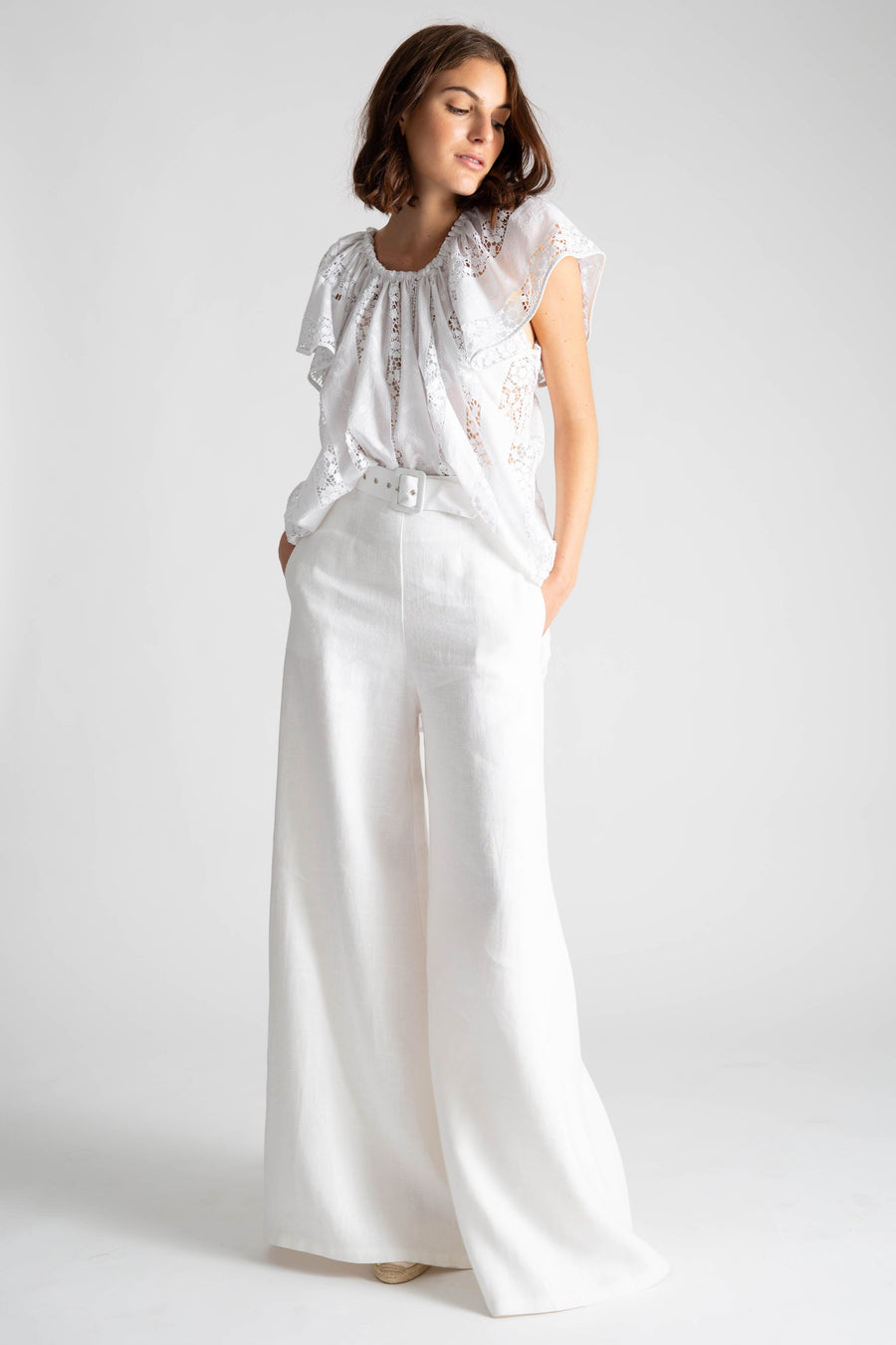 This is a photo of a woman wearing an all white outfit with linen white high waisted flare pants and belt. On top, she wears a high-low top with flowy show sleeves in a cotton lace embroidery.