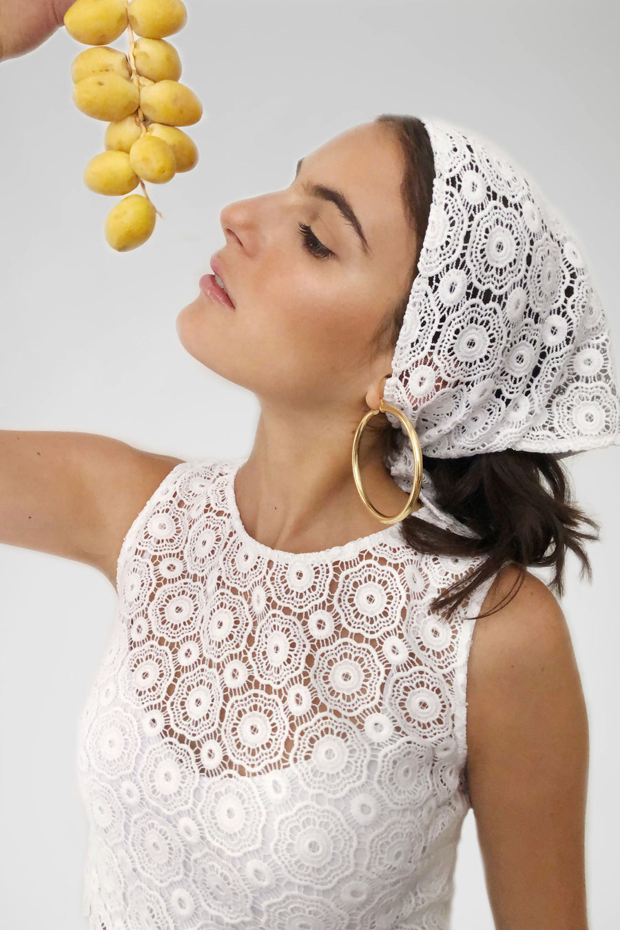 This is a photo of a girl wearing a lace headscarf with gold hoops and matching lace crop top. She is holding fruit above her face.