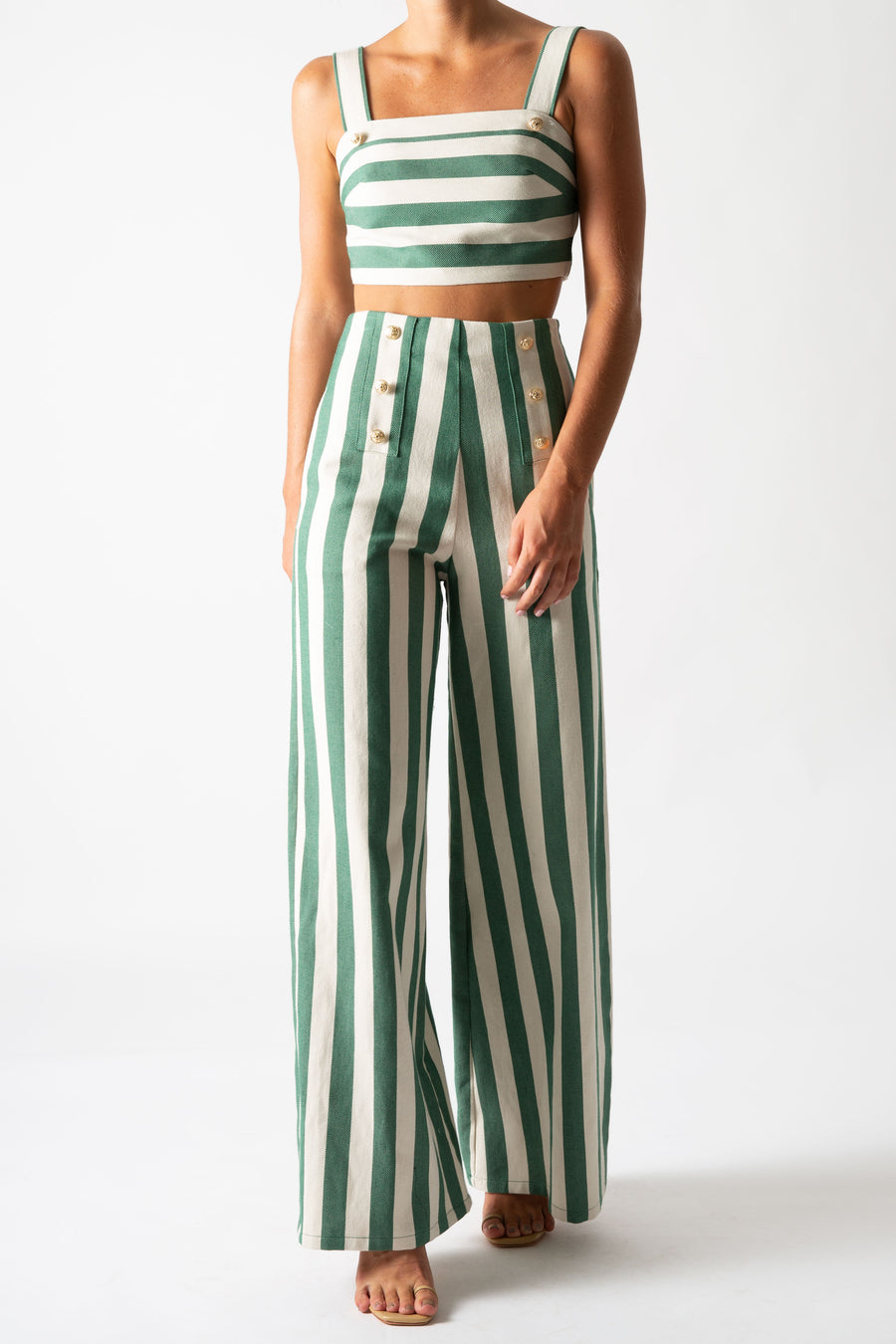 This is a photo of a woman wearing a matching two piece green striped set with gold buttons.  The pants are high waisted and long with a flare and the top is cropped with straps.