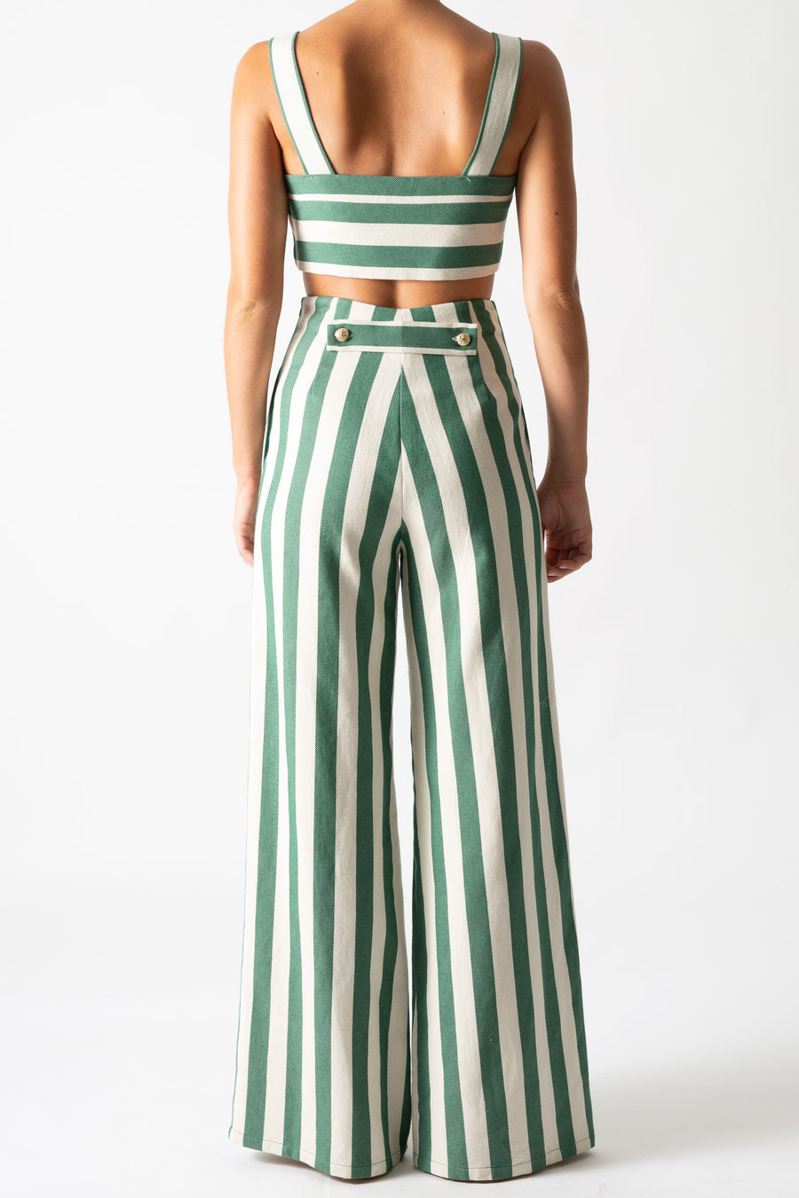 This is a back view photo of a woman wearing a matching two piece green striped set with gold buttons.  The pants are high waisted and long with a flare and the top is cropped with straps.