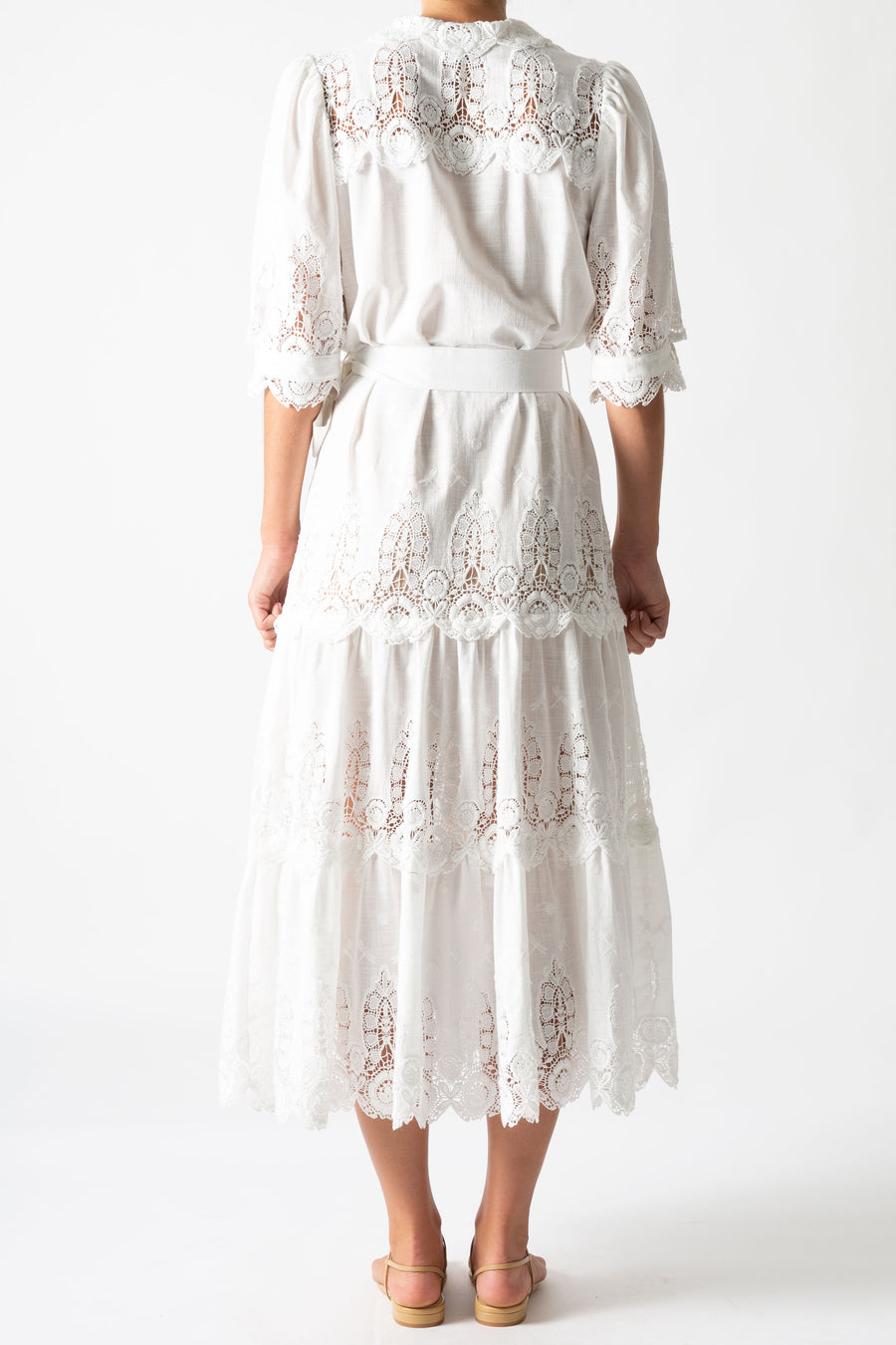This is a back view photo of a woman wearing a long white linen and lace maxi dress with bow tie at the waist and puffy short sleeves.