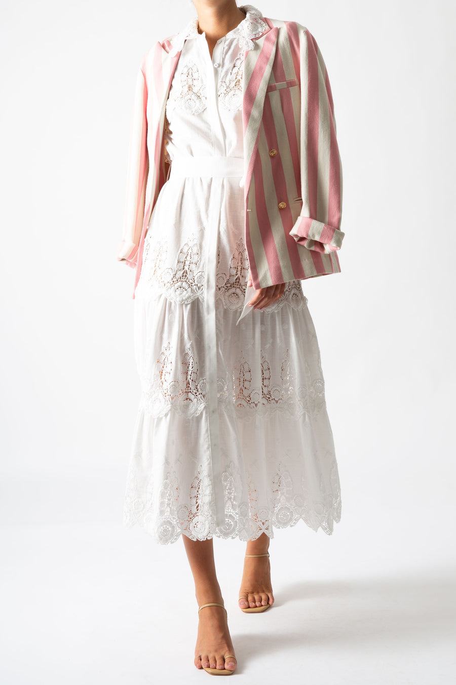 This is a photo of a woman wearing a long white linen and lace maxi dress with bow tie at the waist and puffy short sleeves. She styles it with a pink striped blazer on top.