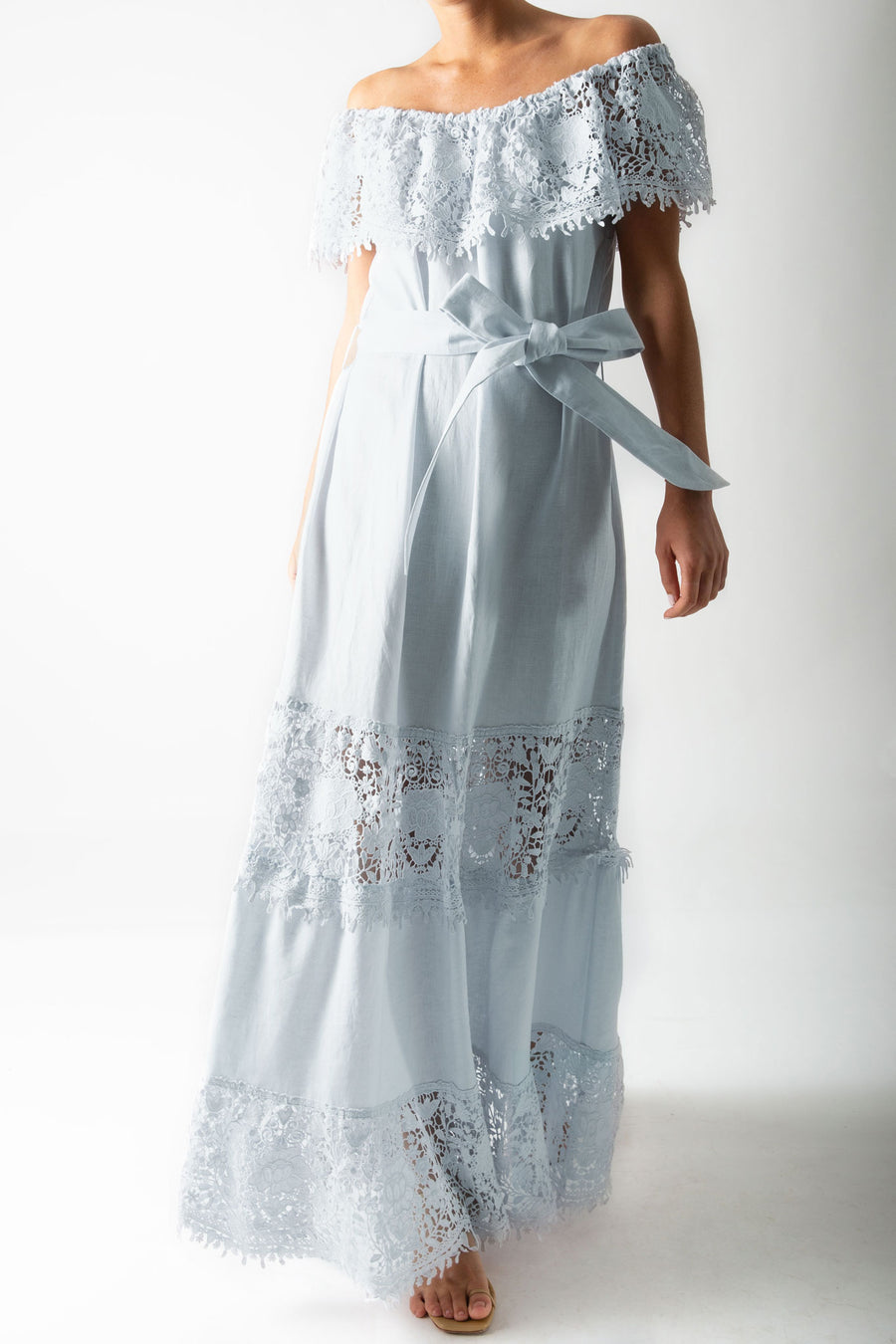 This is a photo of a woman wearing a light blue maxi dress with lace trim across the shoulders, a linen sash for a belt, and tiers of linen and lace down the dress skirt.