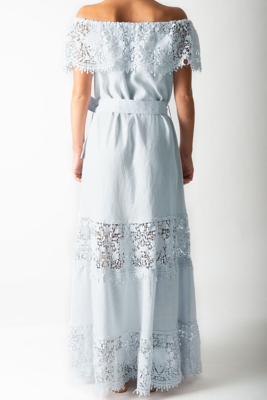 This is a back view photo of a woman wearing a light blue maxi dress with lace trim across the shoulders, a linen sash for a belt, and tiers of linen and lace down the dress skirt.