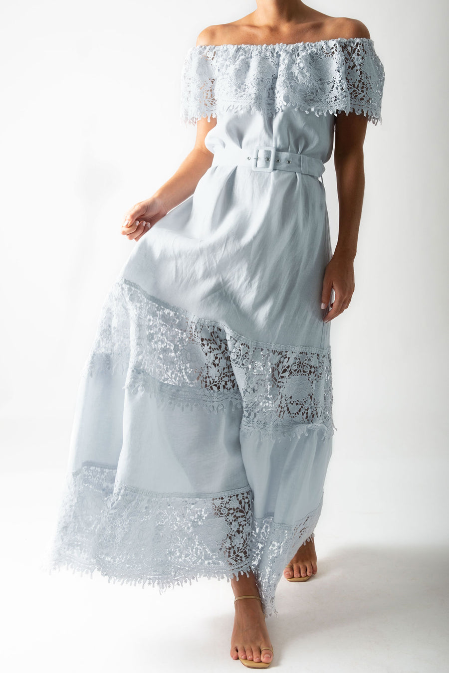 This is a photo of a woman wearing a light blue maxi dress with lace trim across the shoulders, a buckle belt, and tiers of linen and lace down the dress skirt.