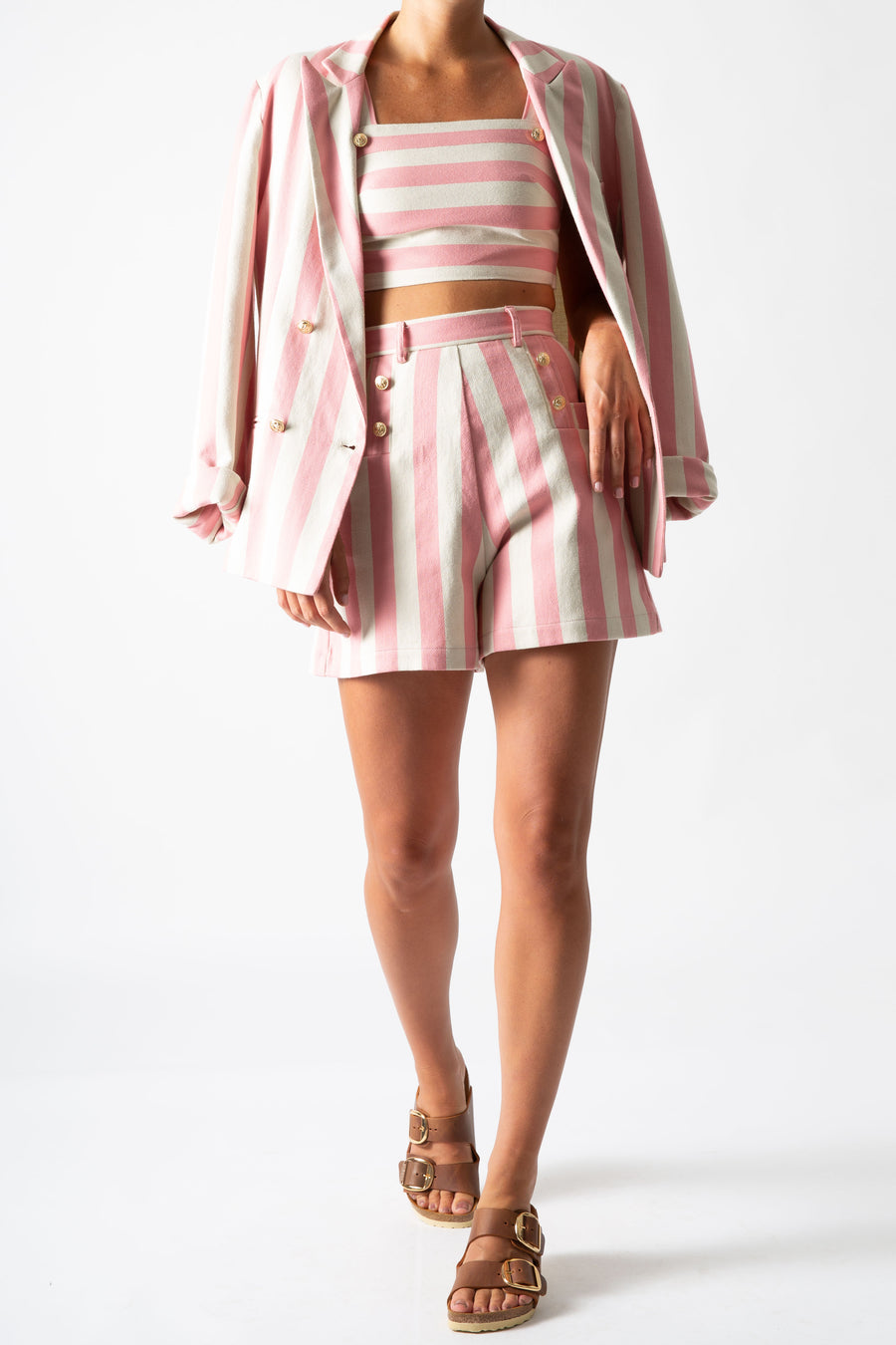 This is a photo of a woman wearing a 3 piece suit with shorts, a cropped top, and blazer over her shoulders. The suit is pink and neutral thick stripes with gold buttons.