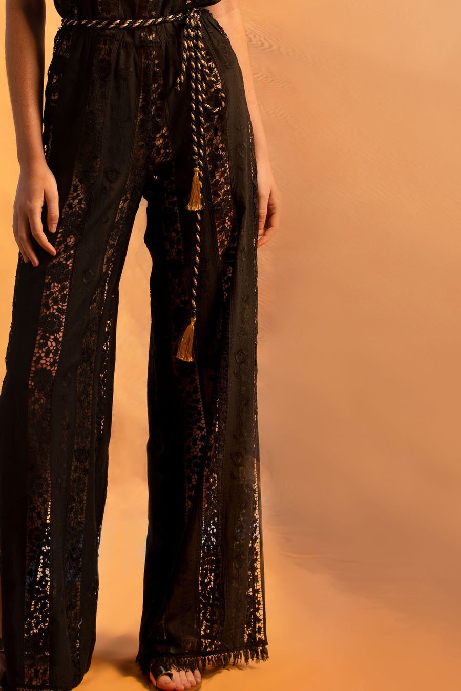 This is a detail photo of the bottom of a black cotton lace embroidered jumpsuit with black fringe on the bottom hemline and side leg slits.