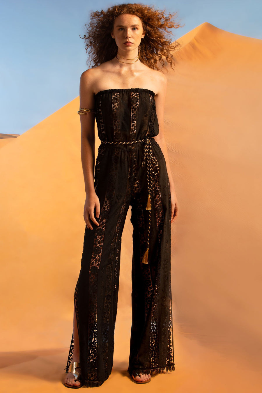 This is a photo of a woman wearing a strapless black cotton and lace embroidered jumpsuit. It has an elastic waistline and side leg slits. The hemline is finished with black fringe and the model wears a gold and black metallic belt around the waist.