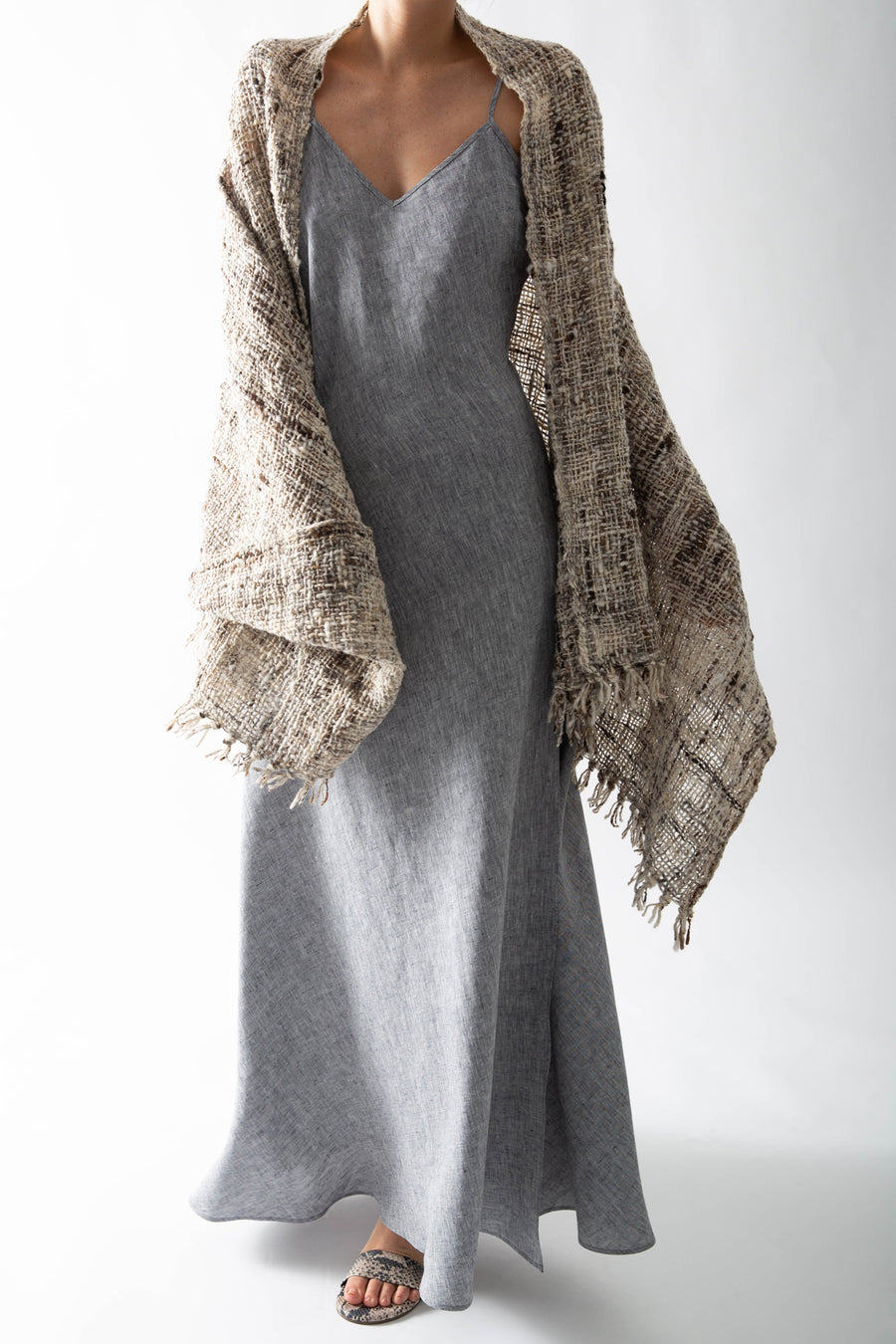This is a photo of a woman from the neck down. She is wearing a long, woven shawl on top of a linen slip dress. The shawl is multicolored, featuring natural shades of gray, brown, and tan and has fringed tassels along the edges.