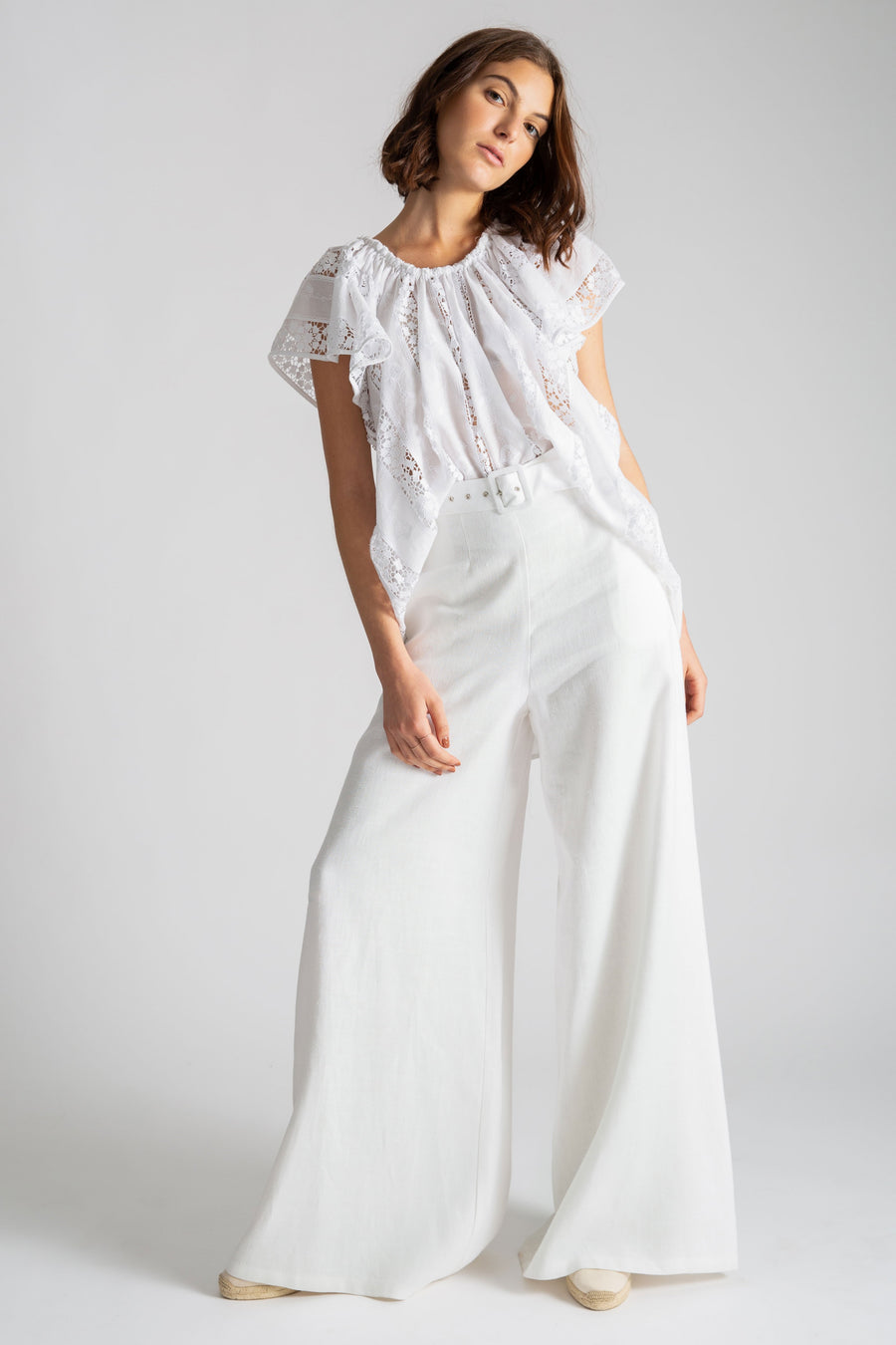 This is a photo of a woman wearing an all white outfit with linen white high waisted flare pants and belt. On top, she wears a high-low top with flowy show sleeves in a cotton lace embroidery.