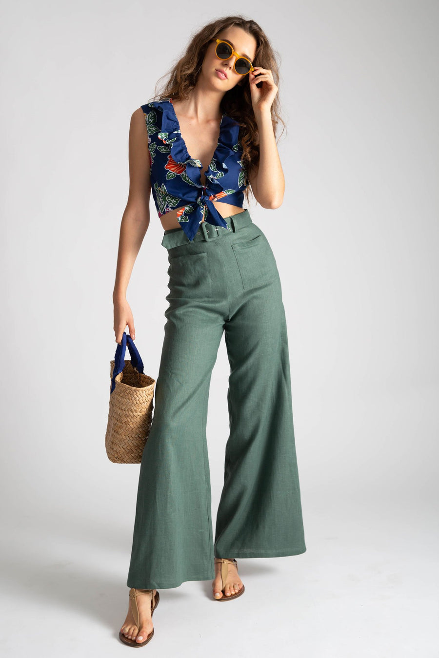 This is a photo of a woman wearing green linen high waisted flare pants with 2 front square pockets and a removable belt. She wears the pants with a floral tie crop top with front ruffle along the deep v neckline. The full look is styled with a natural colored straw bag and yellow sunglasses.