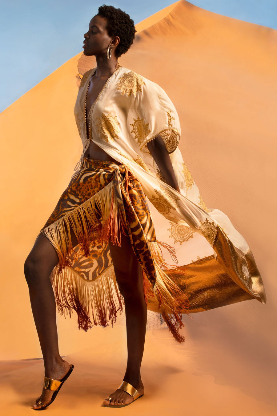 This is a photo of a woman wearing a cheetah pareo with ombre fringe on the hemline. On top, she wears a white coat with gold embellishments and a gold hemline. She stands in front of a desert sand dune.