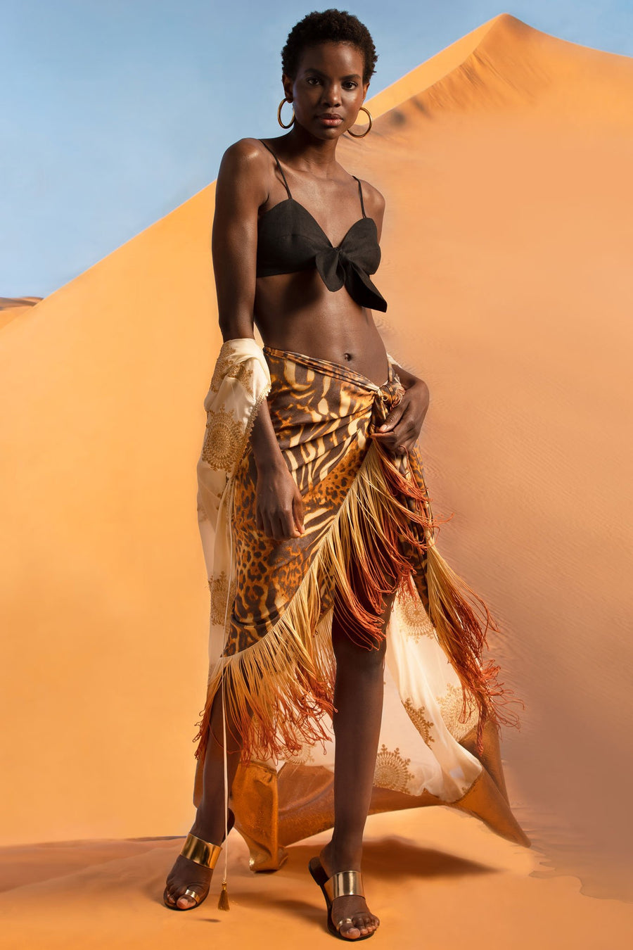 This is a photo of a woman wearing a black linen bralette with center tie, paired with a cheetah pareo with ombre fringe on the hemline. Falling around her arms, she wears a white coat with gold embellishments and a gold hemline. She stands in front of a desert sand dune.