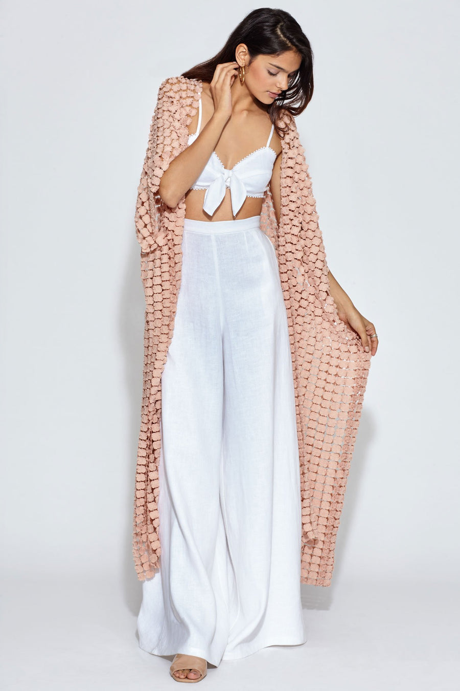 This is a photo of a woman wearing a white linen bralette with white linen wide leg pants. On top of the bralette and pants, she is wearing a dusty pink, dandelion lace cover-up.
