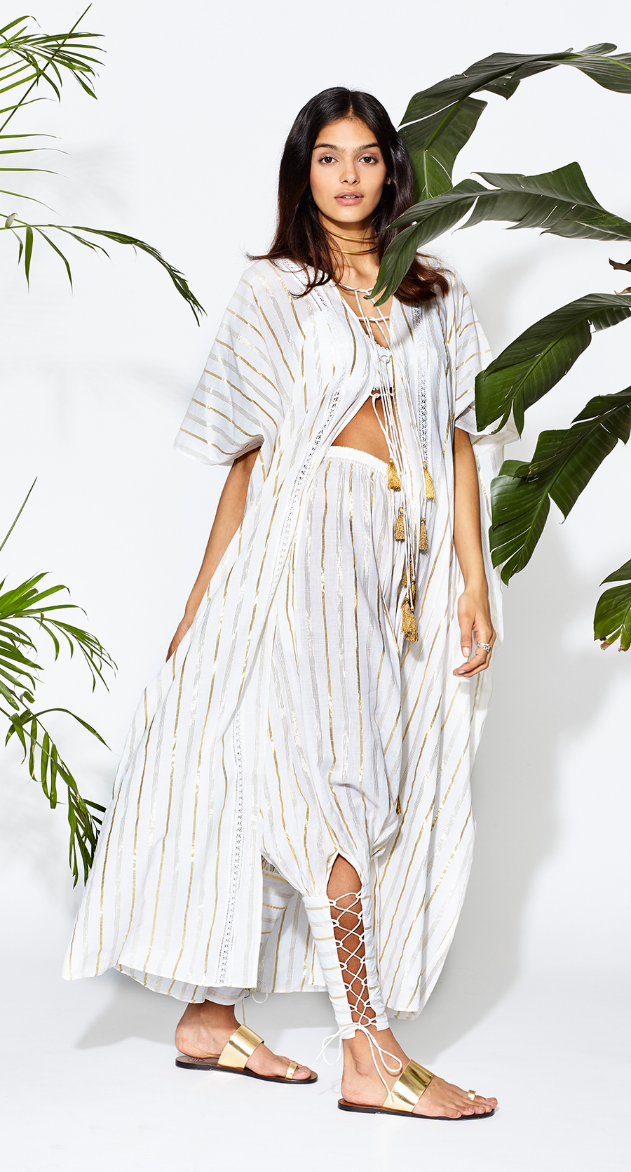 This is a photo of a woman wearing a white coat with silver and gold stripes. She pairs this with a matching pants, a white bralette and gold sandals.