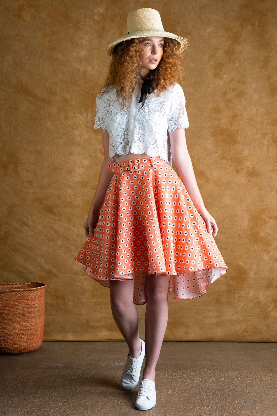 This is a photo of a woman wearing a button up white lace top with a high waisted daisy printed high low midi skirt in coral color. She wears white sneakers and straw safari hat tied around her neck with black ribbon.