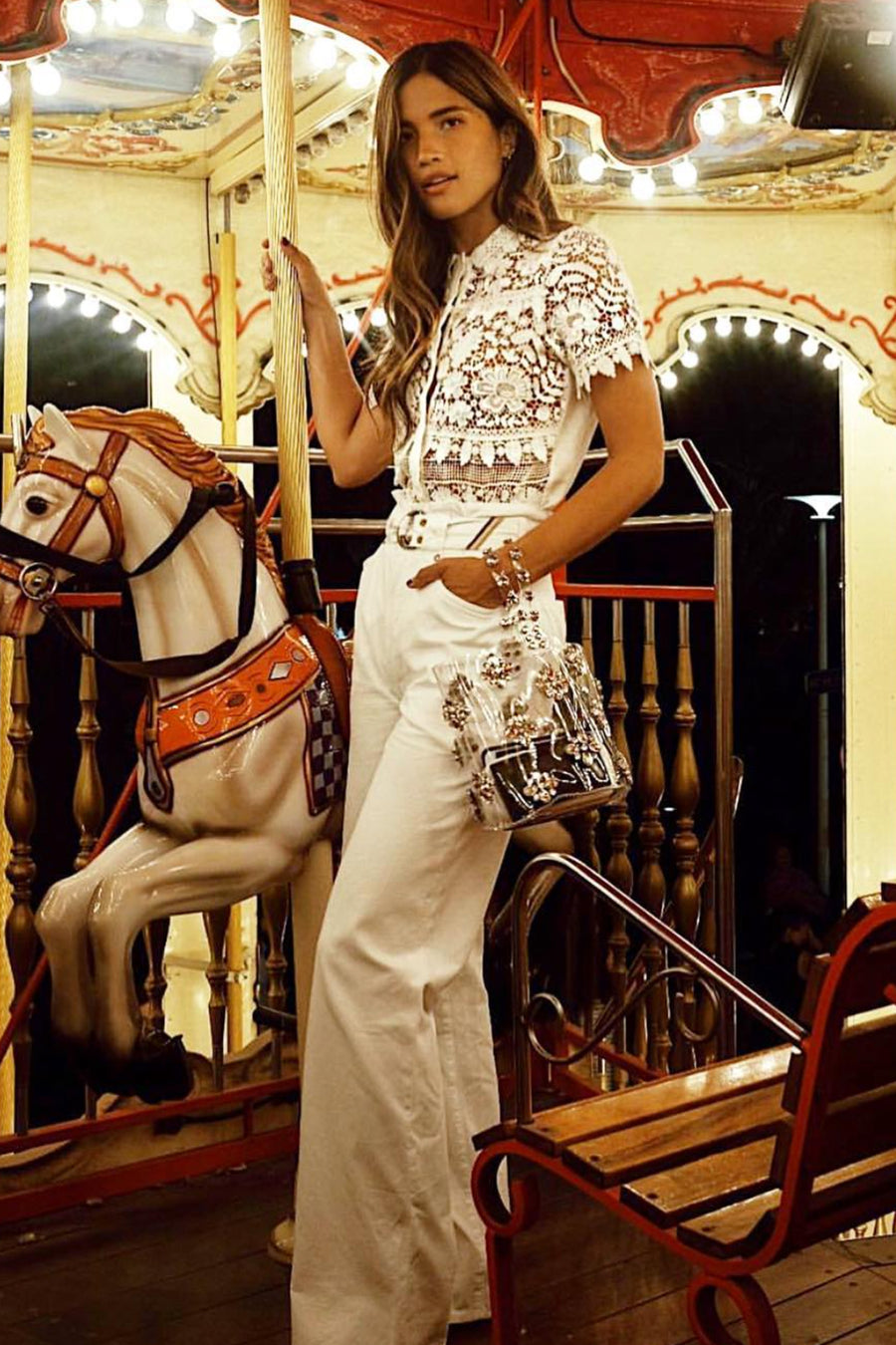 This is a photo of a woman. She is wearing a button front, lace tired blouse tucked into a pair of white, flare leg pants. She has her hand in the left side pocket of the pants.