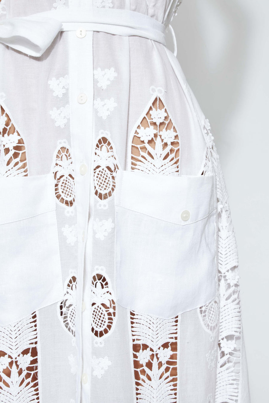 This is a detail photo of square pockets on the front of a white cotton embroidered dress. The pockets fall below the waist and lace design shows small flowers and pineapples.