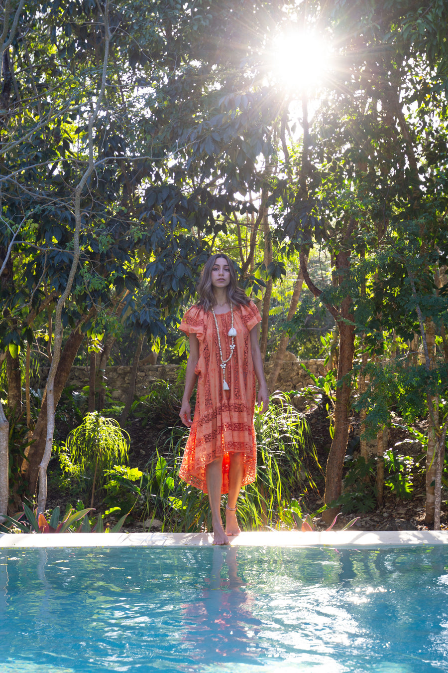 This is a photo of a woman wearing an orange cover up. She stands on the edge of a pool with greenery behind her.