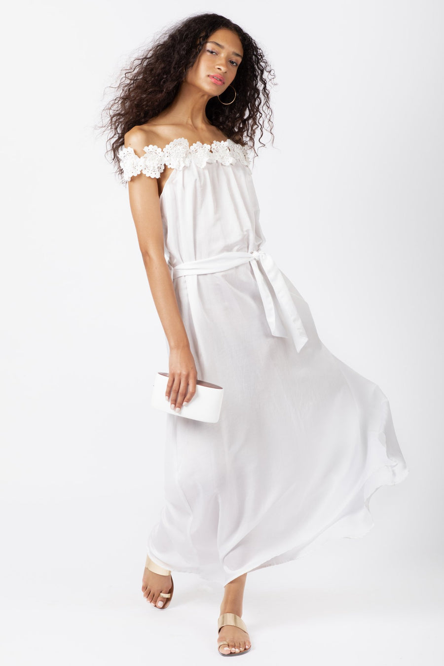 This is a photo of a woman wearing an off the shoulder, white, floor length dress, that has embroidered flower trim all around the neckline.